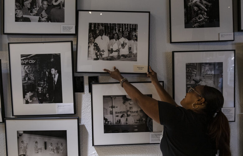 A woman with a long black ponytail reaches up to straighten the frame of one of many black and white photographs displayed in a closely spaced array on a wall in an art gallery.