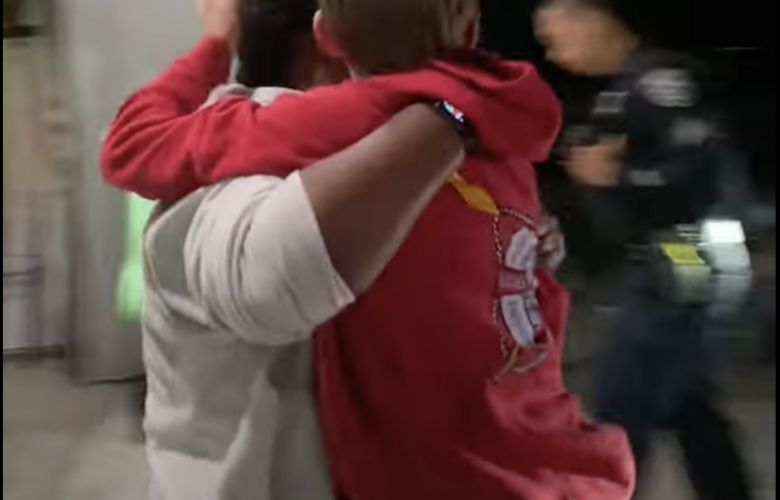 An adult forcibly picks up a boy wearing a red sweatshirt with a hood. Their faces are turned away. A police officer in uniform walks in the background.